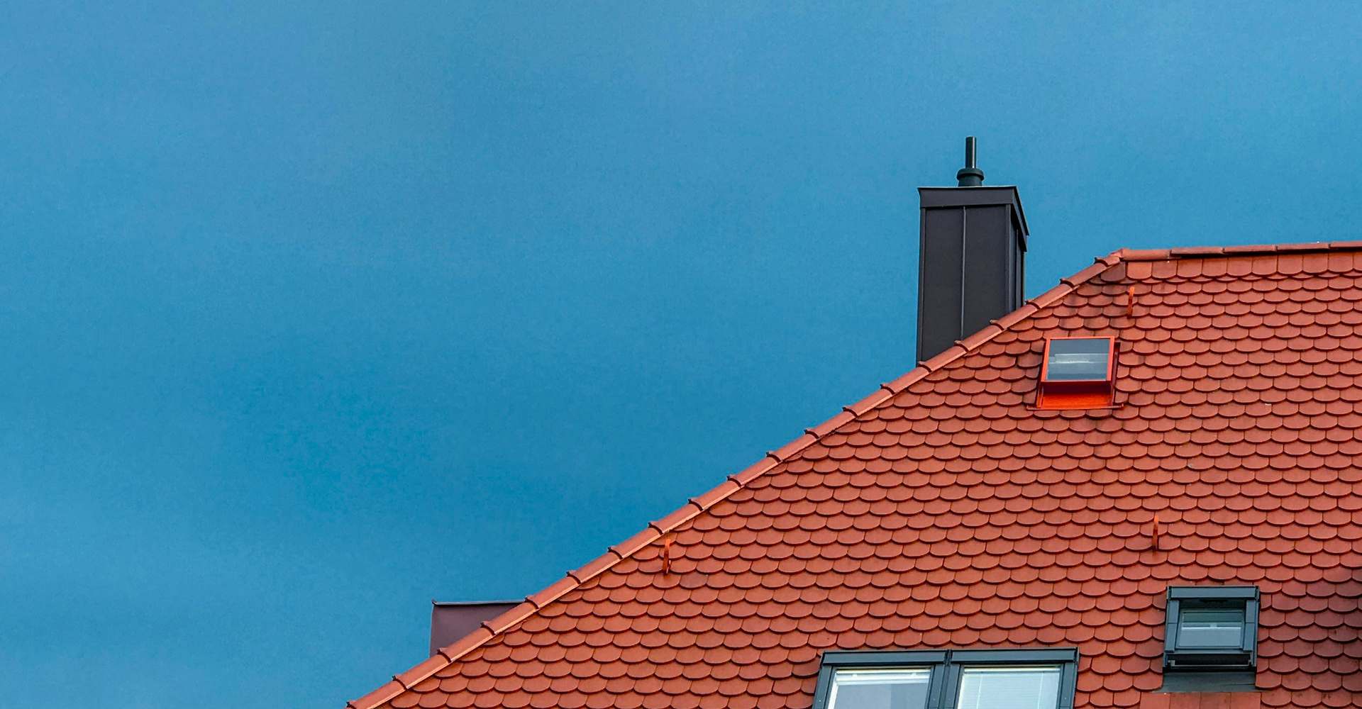 A clay tile roof against a clear blue sky, with a chimney and two skylights visible along the slope.